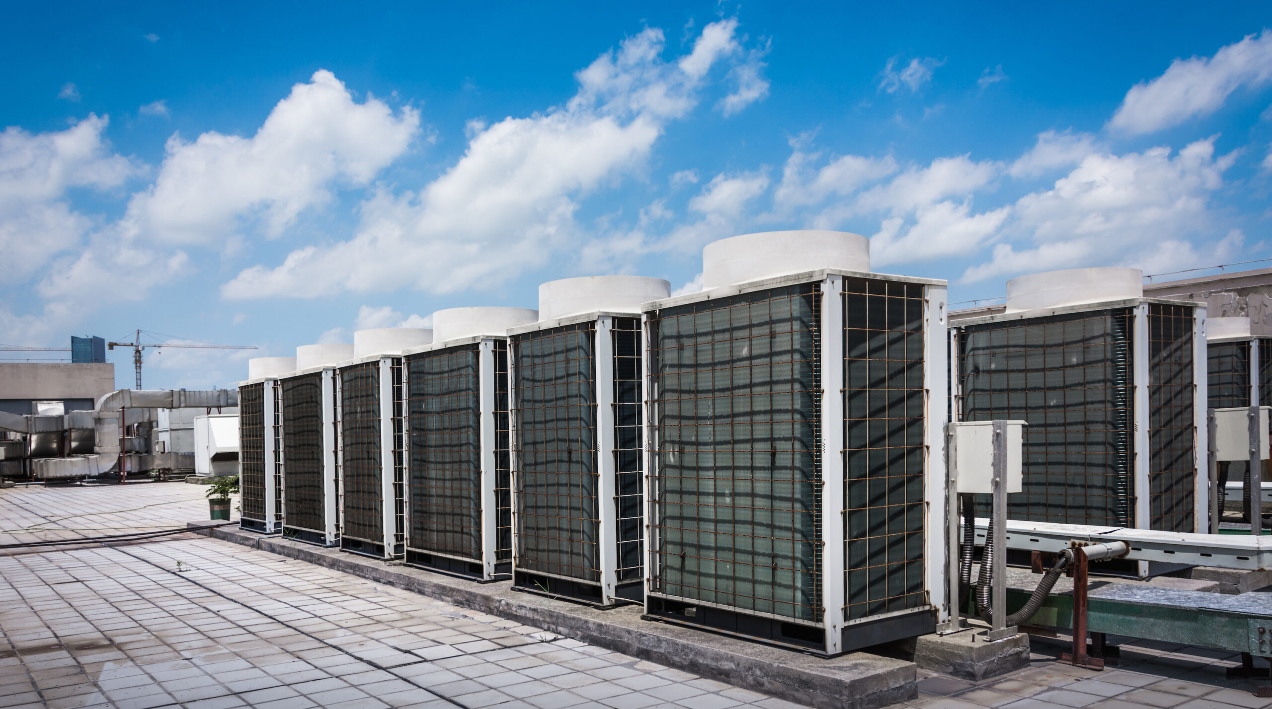 Square Air Conditioning Unit On The Roof With A Round Fan. In The Background Gradually Receding Other Units That Are Out Of Focus. On The Right Side Light Blue Sky And Commercial Space.
